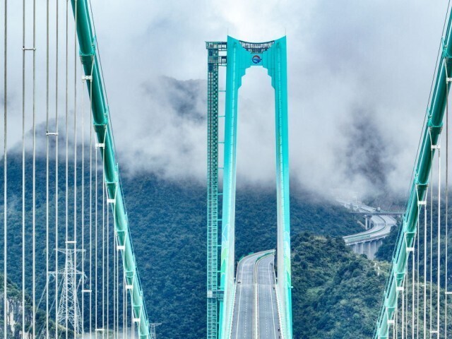 A towering blue suspension bridge with a distinctive curved arch spans across lush, fog-covered mountains, showcasing its height as part of the Guizhou Huajiang Gorge Bridge, the world's tallest bridge.